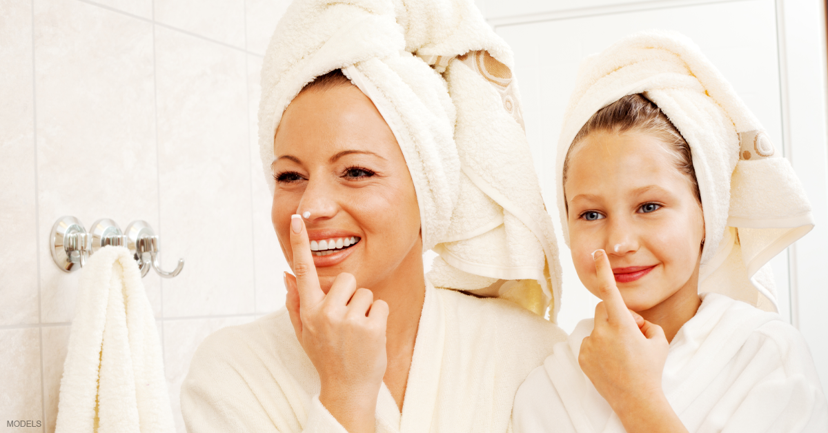 Mom and young daughter smiling and looking in mirror as both dab lotion on their noses