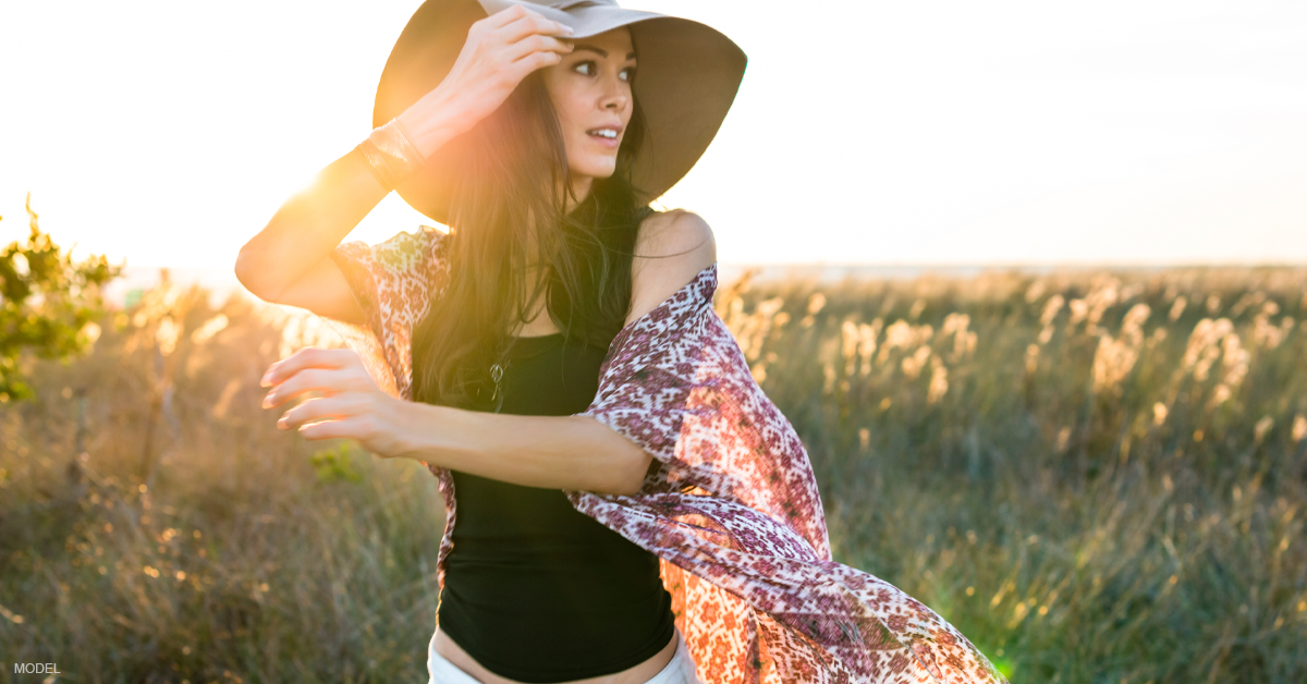 woman in field wearing floppy hat as sun sets behind her